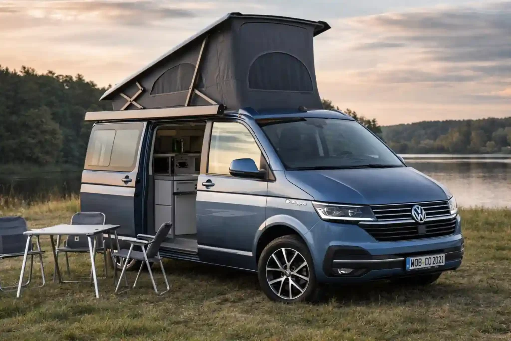 Volkswagen California avec toit relevable ouvert et aménagement camping stationné au bord d’un lac avec table et chaises de camping devant le van aménagé