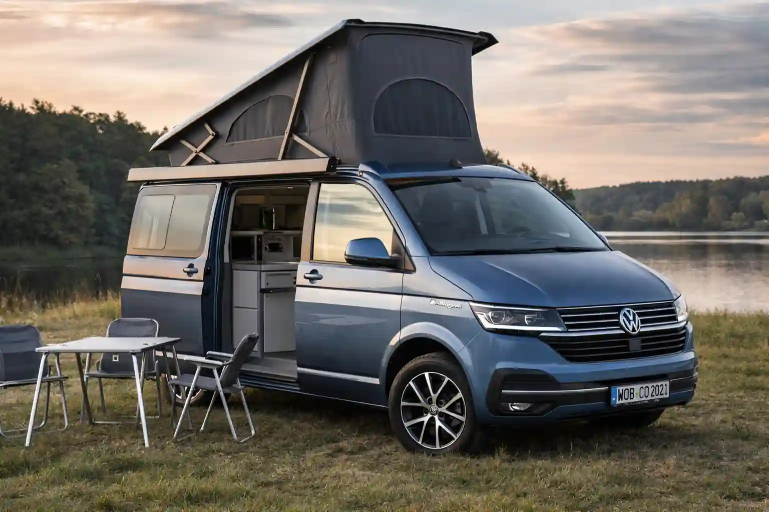 Volkswagen California avec toit relevable ouvert et aménagement camping stationné au bord d’un lac avec table et chaises de camping devant le van aménagé